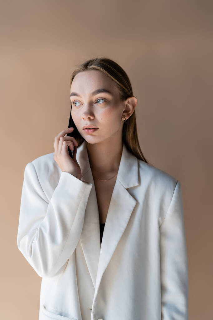 mujer joven en blazer blanco mirando hacia otro lado mientras habla en el teléfono inteligente aislado en beige - Foto, imagen