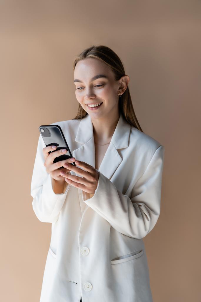 feliz joven en blanco blazer mensajería en el teléfono móvil aislado en beige - Foto, Imagen