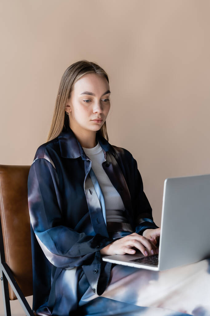 junge Frau in stylischem Oversize-Hemd sitzt und tippt auf Laptop isoliert auf beige - Foto, Bild