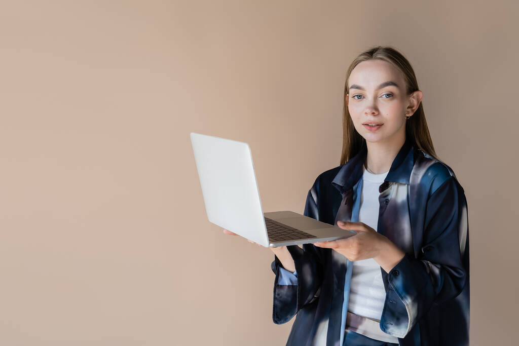 woman in stylish oversize shirt looking at camera while holding laptop isolated on beige - Foto, immagini