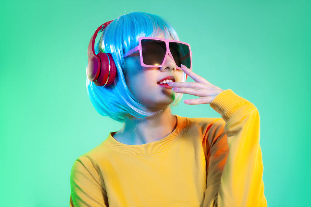 Junges asiatisches Mädchen in gelbem Sweatshirt mit blauer Schnurrfrisur, rotem Kopfhörer und rosa Sonnenbrille hört Musik, die auf dem Hintergrund des grünen Bildschirms posiert. - Foto, Bild