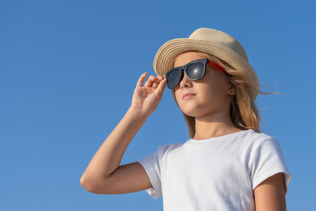 A young girl in a hat and sunglasses adjusts her glasses with one hand and looks to the side. - 写真・画像