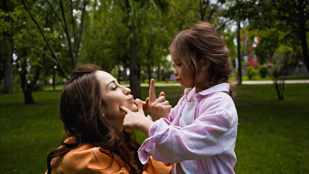 happy mother pouting lips and pointing at nose of cute daughter in green park - Foto, imagen