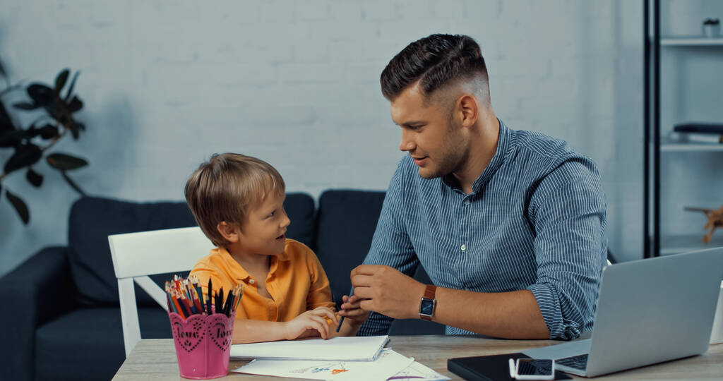 Отец держит карандаш и смотрит на сына возле гаджетов на столе - Фото, изображение