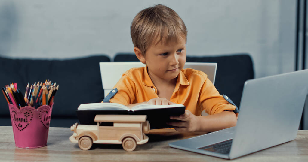 niño sosteniendo portátil y mirando portátil cerca de coche de juguete de madera en el escritorio - Foto, Imagen