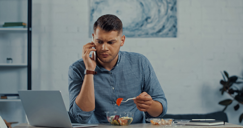 фрілансер тримає пластикову виделку та обідає під час розмови на смартфоні біля ноутбука на столі
 - Фото, зображення