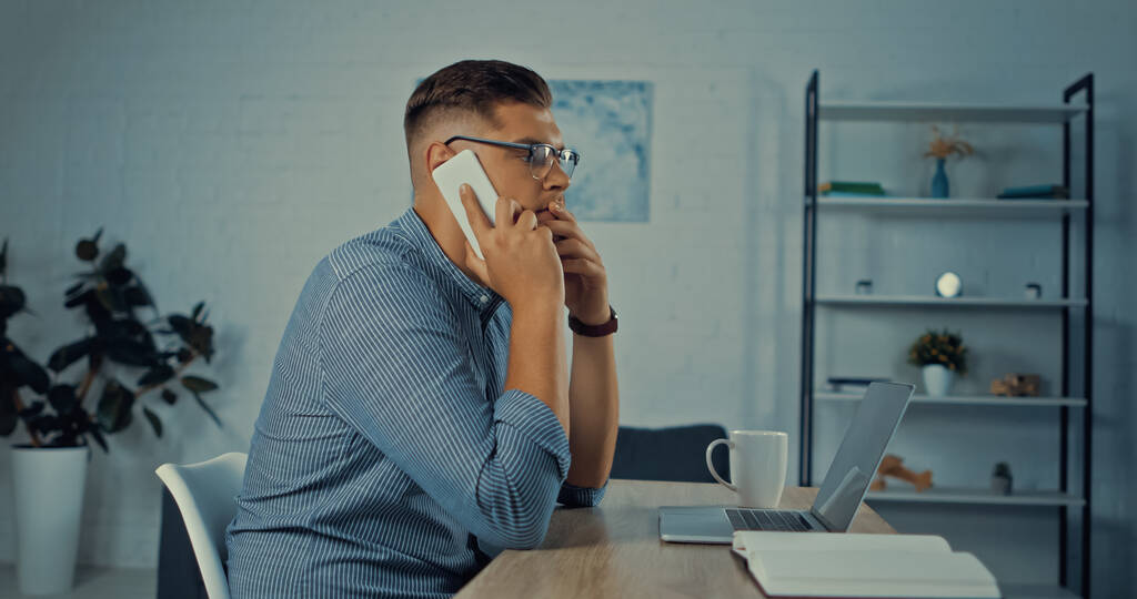 vista lateral del hombre preocupado en gafas hablando en el teléfono inteligente mientras trabaja de forma remota en casa - Foto, imagen