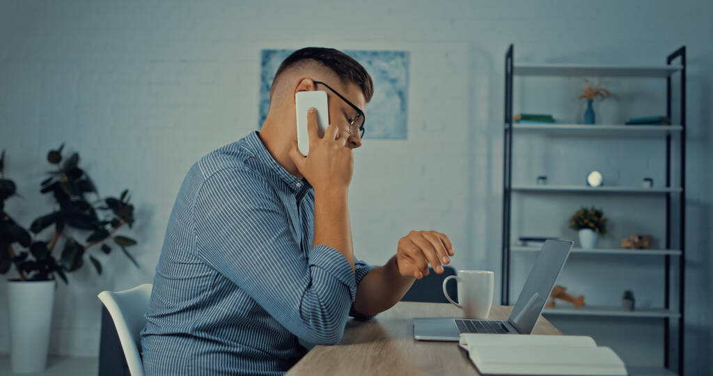 vista lateral do homem de óculos falando no telefone celular enquanto trabalhava remotamente em casa - Foto, Imagem
