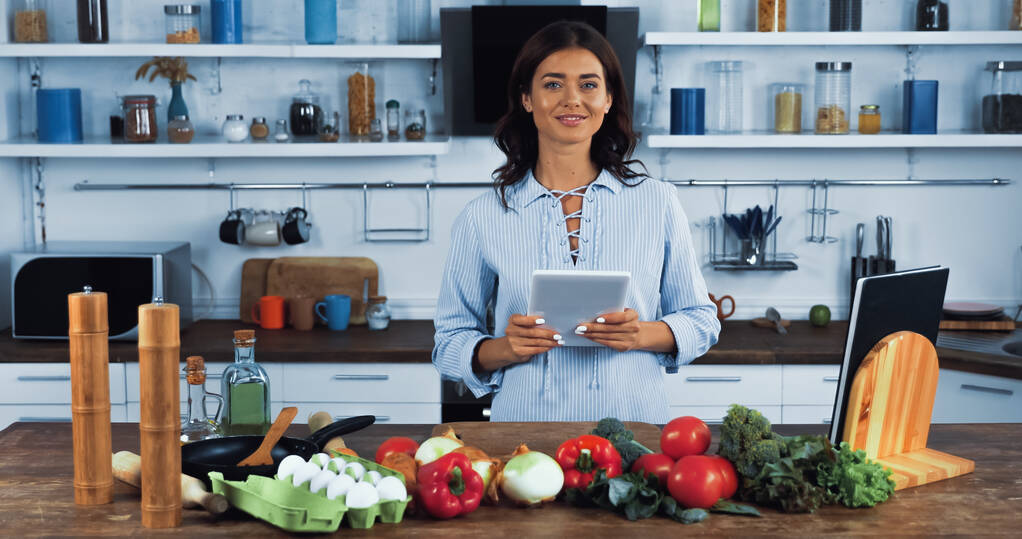 Vegetarierin mit digitalem Tablet lächelt in der Nähe von Rohstoffen in der Küche in die Kamera - Foto, Bild