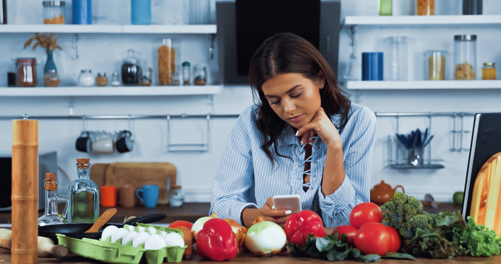 Frau mit Smartphone in der Nähe verschiedener Zutaten und Kochbuch auf Küchentisch - Foto, Bild