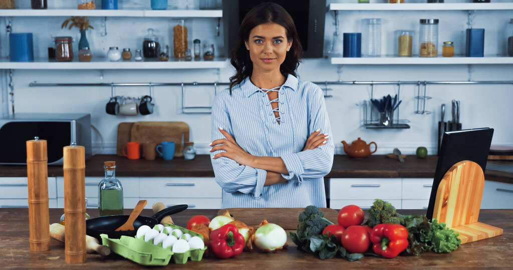 brunette femme avec les bras croisés souriant à la caméra près de différents légumes et œufs  - Photo, image