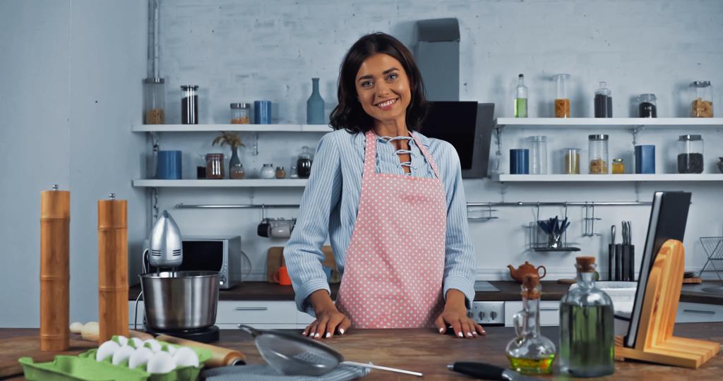 szczęśliwa kobieta w fartuchu w pobliżu procesora żywności i składników na stole kuchennym - Zdjęcie, obraz