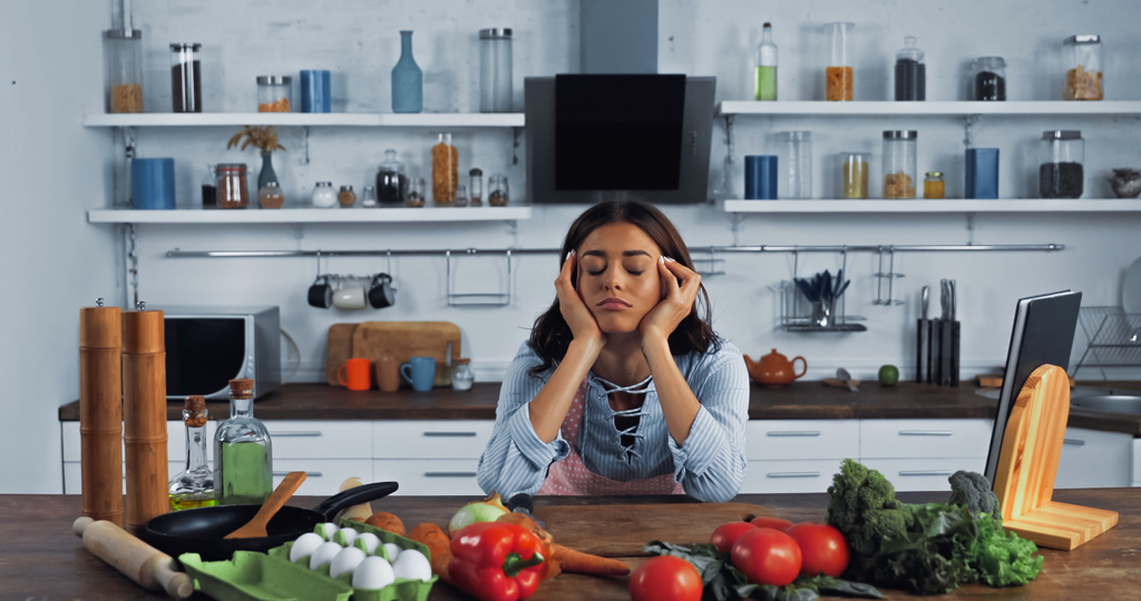 уставшая женщина с закрытыми глазами рядом с сырыми ингредиентами и кухонной утварью на кухне - Фото, изображение