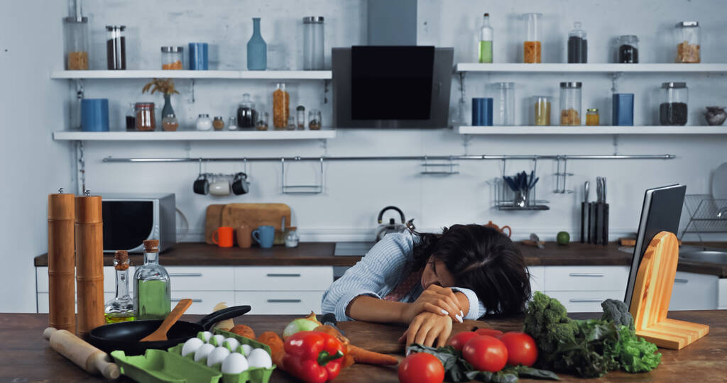 vermoeide vrouw ligt in de buurt van verse groenten en eieren op keukenblad - Foto, afbeelding