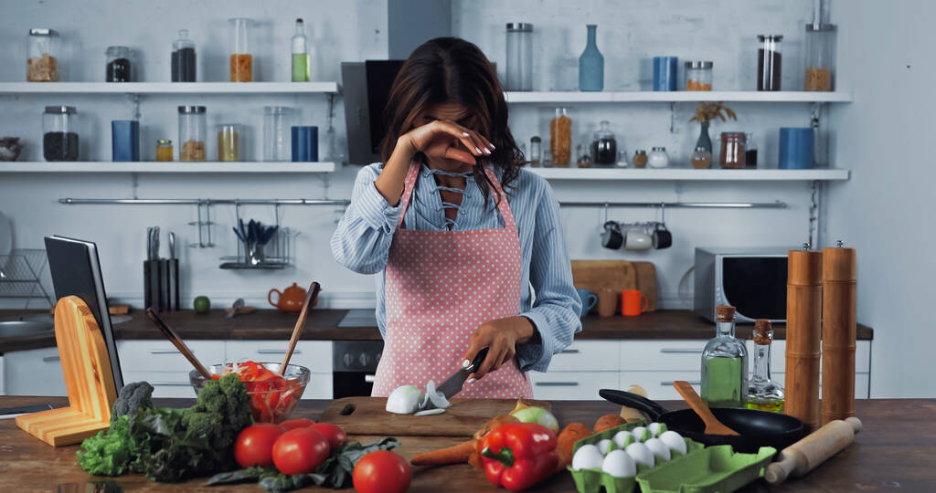 woman crying and wiping irritated eyes while cutting onion near fresh vegetables - Foto, afbeelding