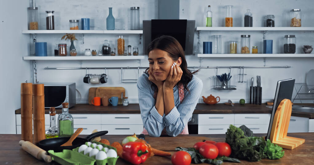 brunette woman smiling near fresh vegetables and cooking utensils on kitchen worktop - Φωτογραφία, εικόνα