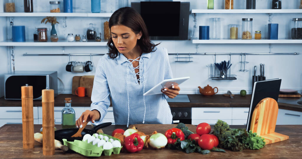 brunette vrouw met digitale tablet bereiken van rauwe eieren in de buurt van verse groenten op werkblad - Foto, afbeelding