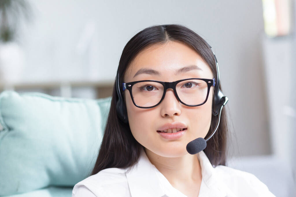 Portret van een jonge Aziatische vrouw in een koptelefoon met een microfoon. Werkt als consultant in een call center, beantwoordt oproepen. Zittend aan de tafel in een bril aan de computer, kijkend naar de camera, glimlachend. - Foto, afbeelding
