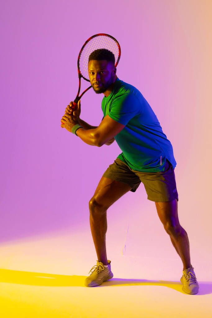 Vertical image of african american male tennis player in violet and yellow neon lighting. Sport, movement, fitness and active lifestyle concept. - 写真・画像