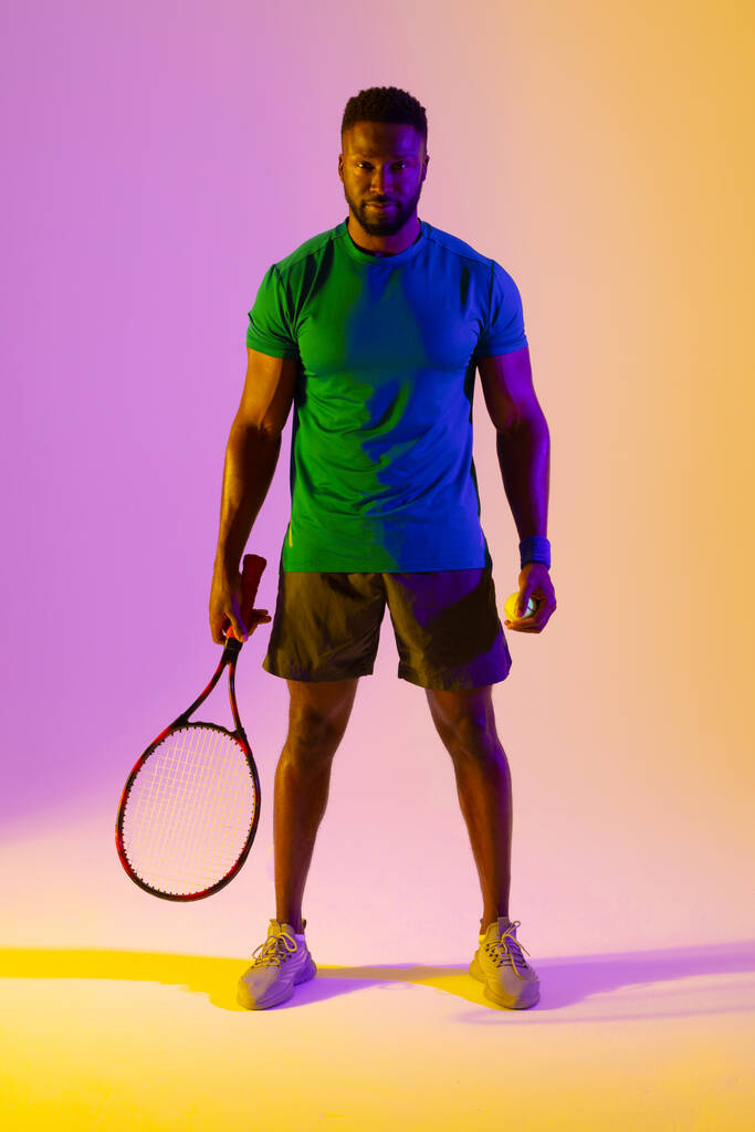 Vertical image of african american male tennis player in violet and yellow neon lighting. Sport, movement, fitness and active lifestyle concept. - Photo, Image