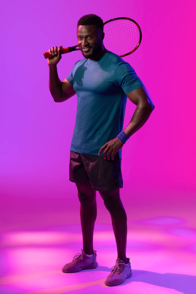 Vertical image of happy african american male tennis player in neon pink lightning. Sport, movement, fitness and active lifestyle concept. - Fotó, kép