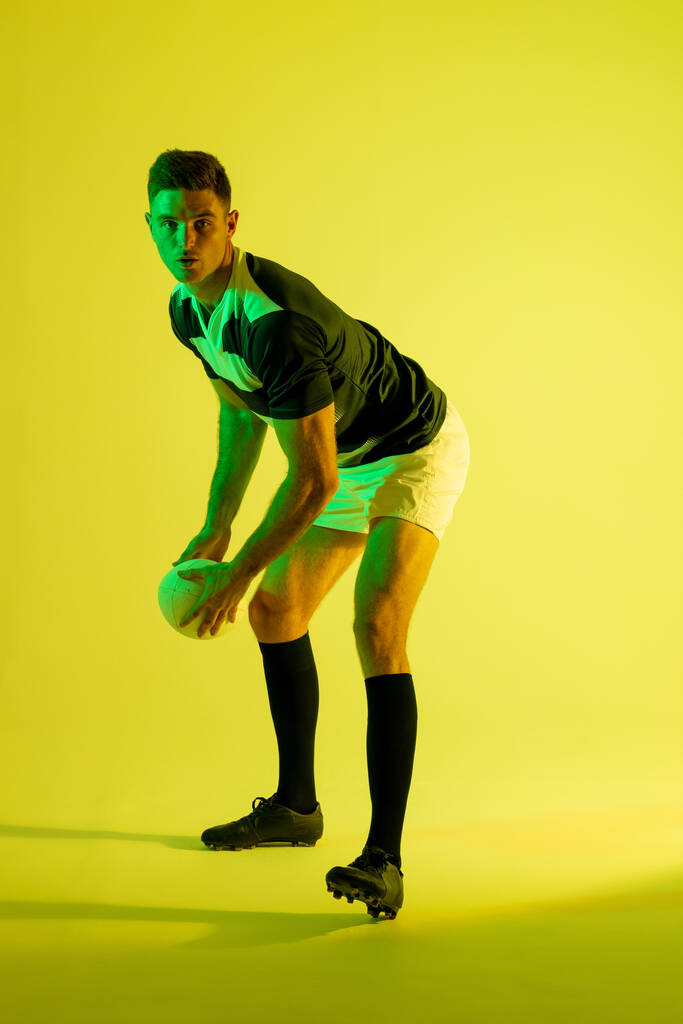 Ritratto di giocatore di rugby maschile caucasico con pallone da rugby su illuminazione gialla. Sport, movimento, allenamento e stile di vita attivo. - Foto, immagini