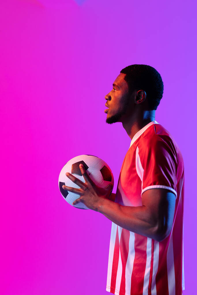 African american male soccer player with football over neon pink lighting. Sport, movement, training and active lifestyle concept. - Photo, Image