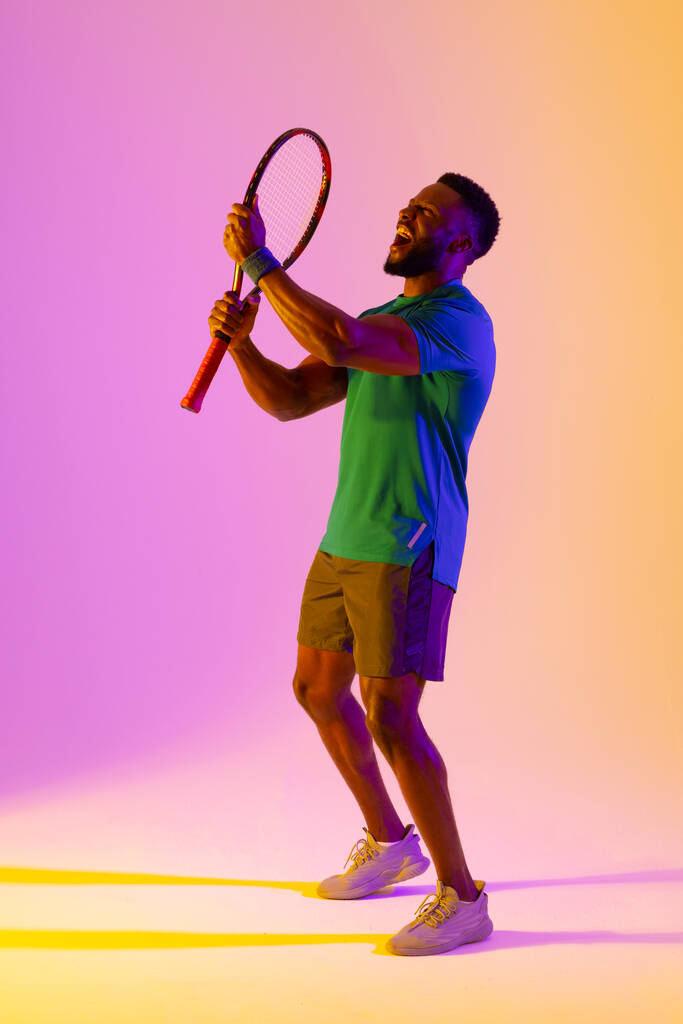 Imagen vertical del exitoso tenista afroamericano masculino en iluminación violeta y amarilla. Deporte, movimiento, fitness y estilo de vida activo. - Foto, Imagen