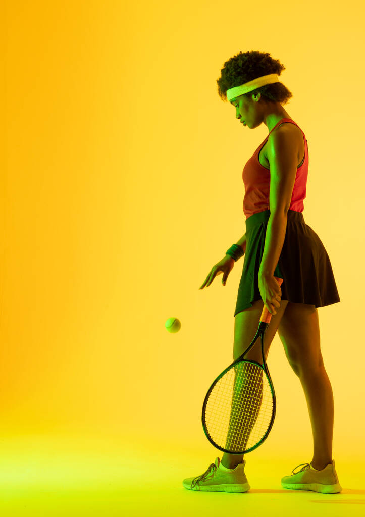 Imagem vertical de uma jogadora de ténis afro-americana em iluminação amarela. Esporte, movimento, fitness e conceito de estilo de vida ativo. - Foto, Imagem
