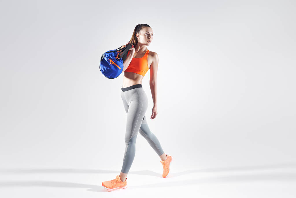 Young woman in sports clothing carrying a bag while walking against white background - Foto, afbeelding