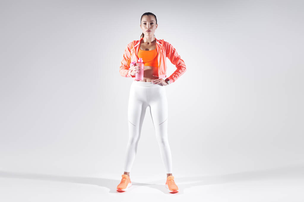 Πλήρες μήκος της όμορφης νεαρής γυναίκας στα αθλητικά ρούχα πόσιμο νερό σε λευκό φόντο - Φωτογραφία, εικόνα