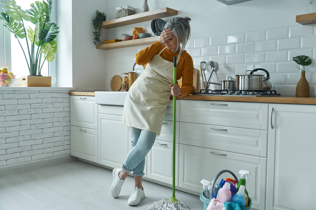Ev mutfağında ayakta dururken paspasa yaslanan yorgun ve yaşlı bir kadın. - Fotoğraf, Görsel