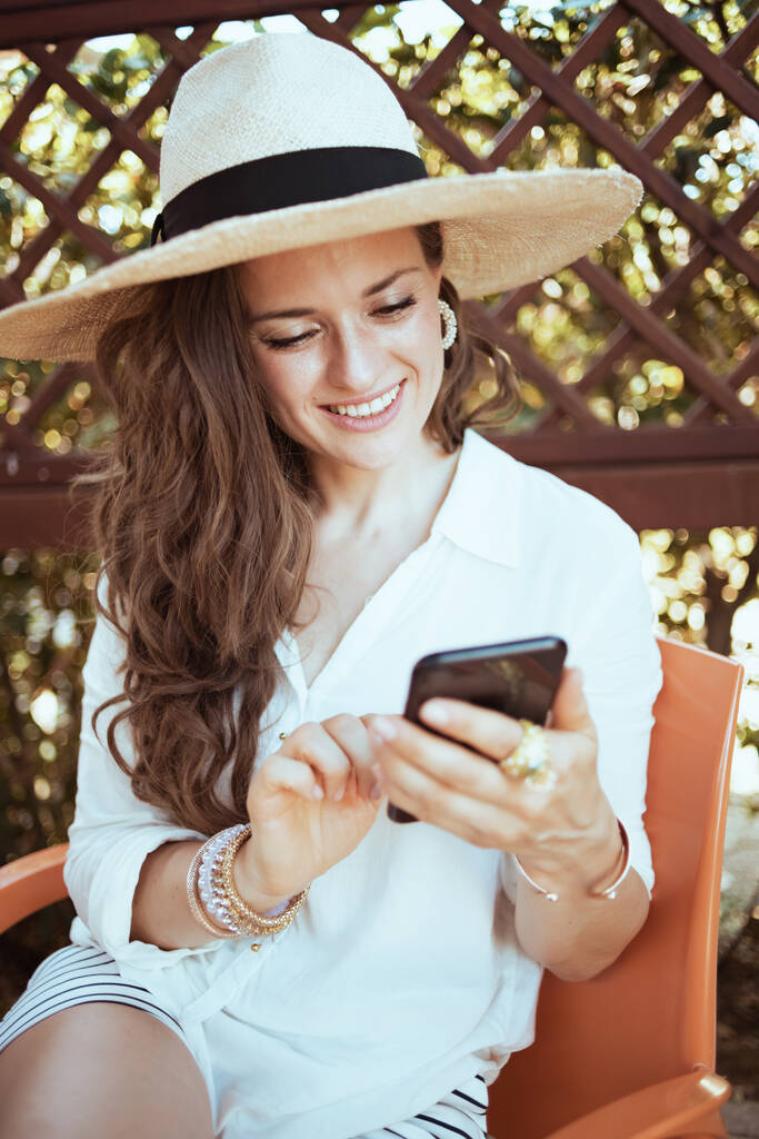 hymyilevä trendikäs nainen valkoisessa paidassa hattu käyttäen älypuhelimen sovelluksia patiolla. - Valokuva, kuva