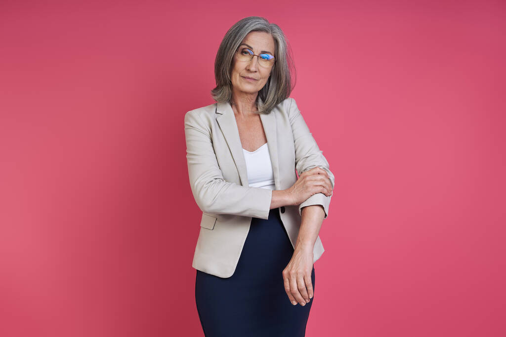 Confident senior woman rolling up sleeve on her jacket while standing against pink background - Photo, Image