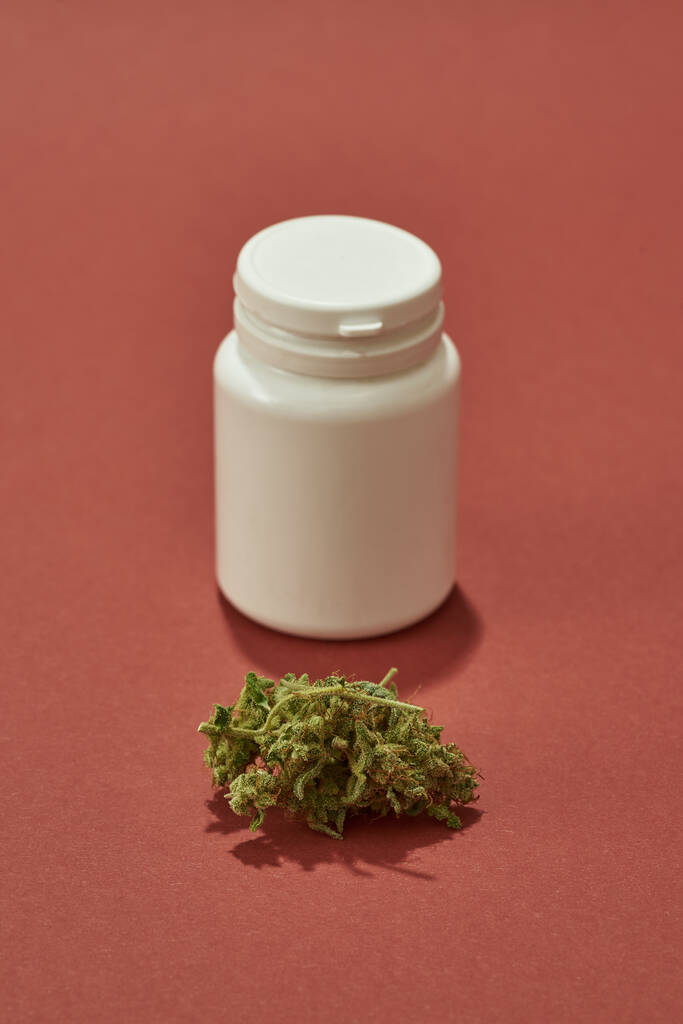 Fresh green dry crushed marijuana weed and plastic jar isolated on red background. Light drug and addiction. Herbal medicine and painkiller therapy. Natural homemade organic cannabis. Copy space - Zdjęcie, obraz