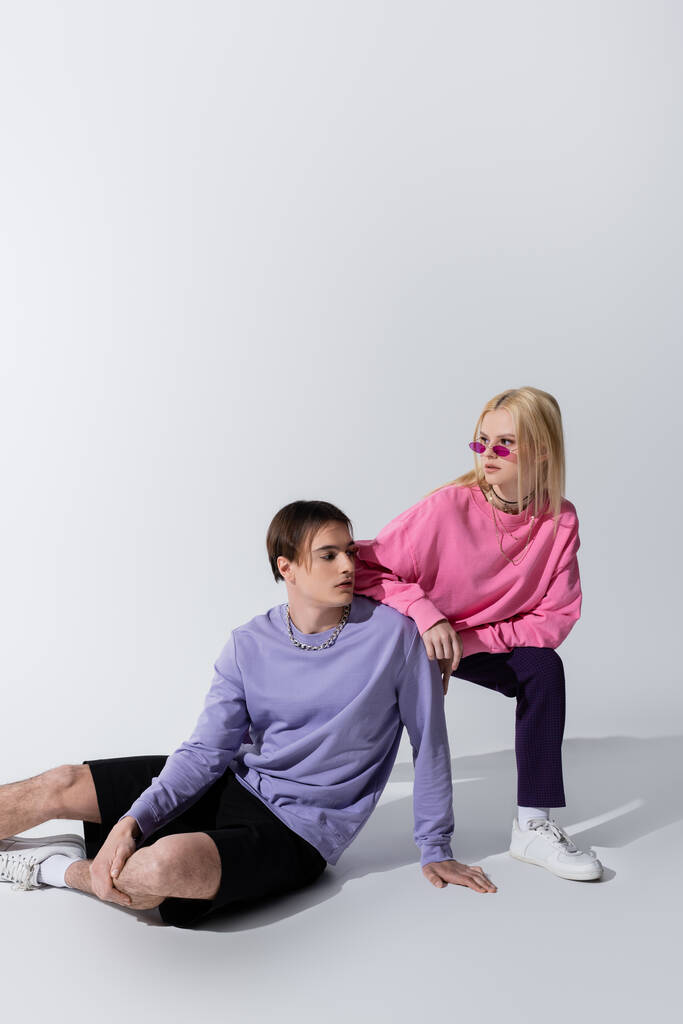 Pretty young model in sunglasses posing near boyfriend sitting on grey background - Foto, afbeelding