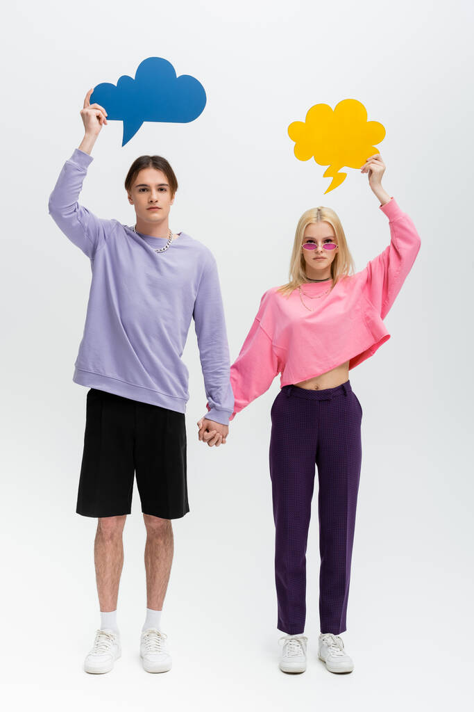 Full length of stylish couple with thought bubbles holding hands on grey background - 写真・画像
