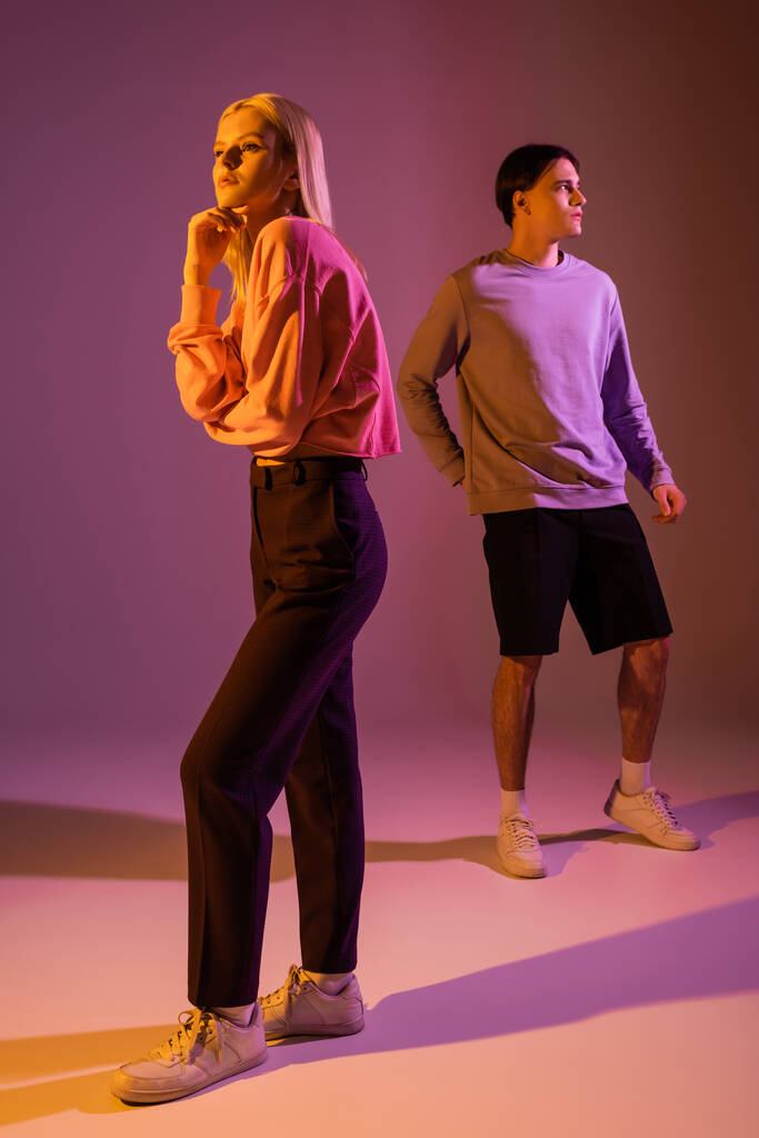 Полная длина красивая блондинка позирует рядом с парнем на фиолетовом фоне с освещением  - Фото, изображение