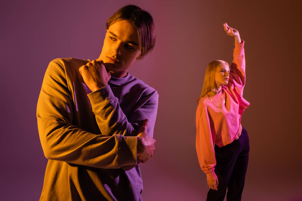 Young brunette man posing near blurred girlfriend on purple background with lighting  - Photo, Image
