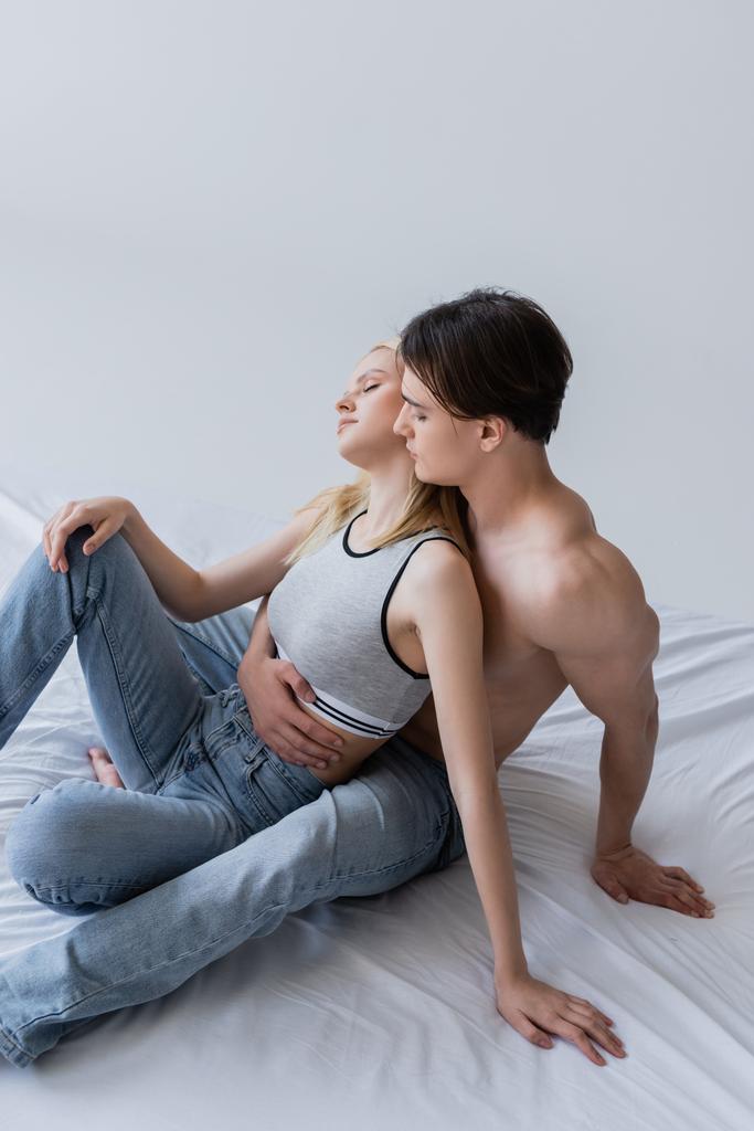 Seksikäs mies syleilee tyttöystävä päälle ja farkut sängyssä eristetty harmaa  - Valokuva, kuva