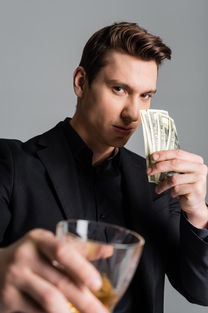 confident man showing dollar banknotes and toasting with blurred glass of whiskey isolated on grey - Photo, Image