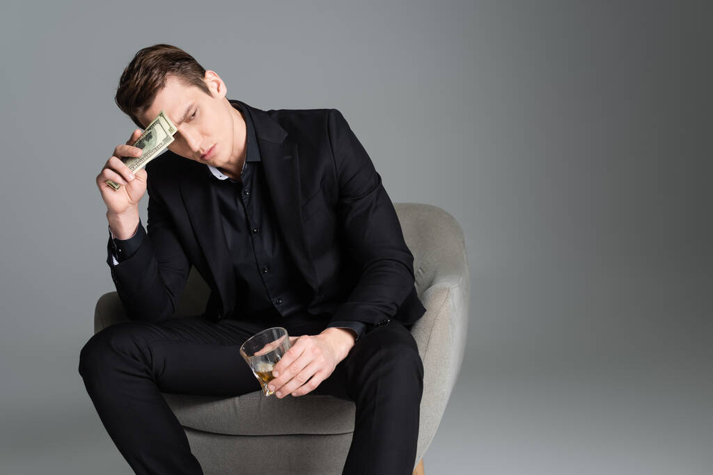 troskliwy człowiek ze szklanką whisky i pieniędzmi siedzący w fotelu i odwracający wzrok na szarym - Zdjęcie, obraz