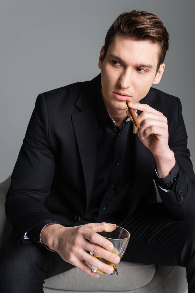 уверенный в себе человек в черном держит сигару и стакан виски, глядя в сторону изолированы на серый - Фото, изображение