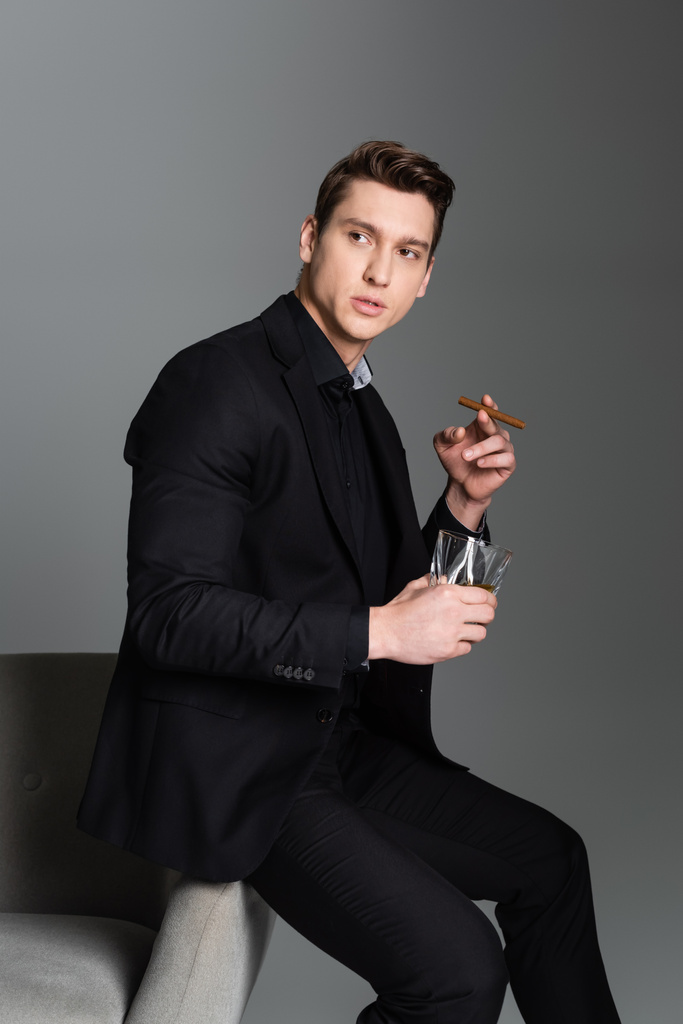 serious man in black blazer looking away while sitting with whiskey and cigar isolated on grey - Fotó, kép