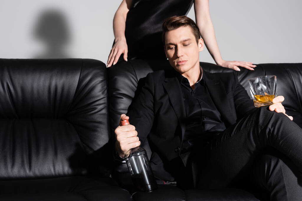 стильный мужчина с бутылкой и стаканом виски на черном диване рядом с женщиной на сером фоне - Фото, изображение