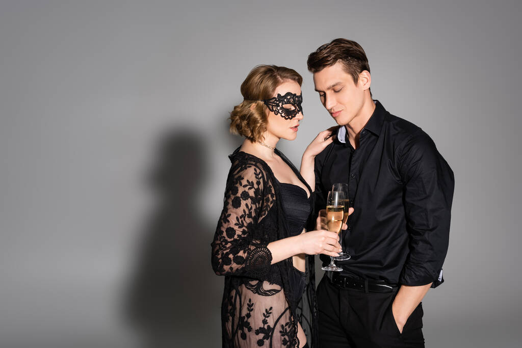 woman in lace mask and tunic clinking champagne glasses with man on grey background - Foto, Bild