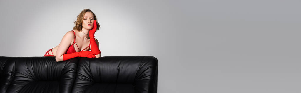 sensuale giovane donna in reggiseno rosso e guanti guardando la fotocamera vicino al divano nero isolato su grigio, banner - Foto, immagini