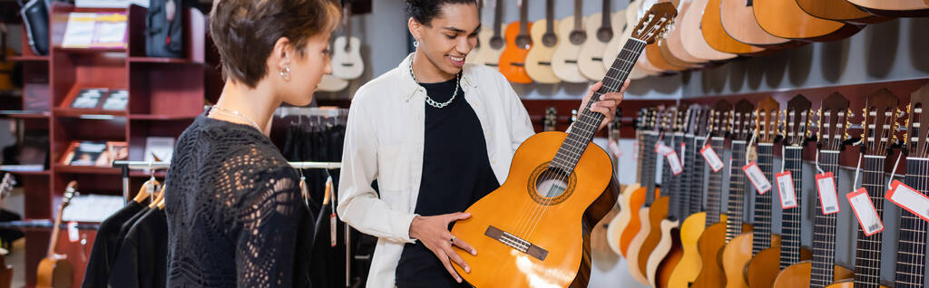 Positive african american seller holding acoustic guitar near woman in music tore, banner  - 写真・画像