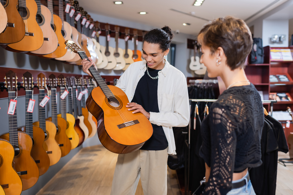 Alegre vendedor afroamericano sosteniendo guitarra acústica cerca de mujer borrosa en tienda de música - Foto, Imagen