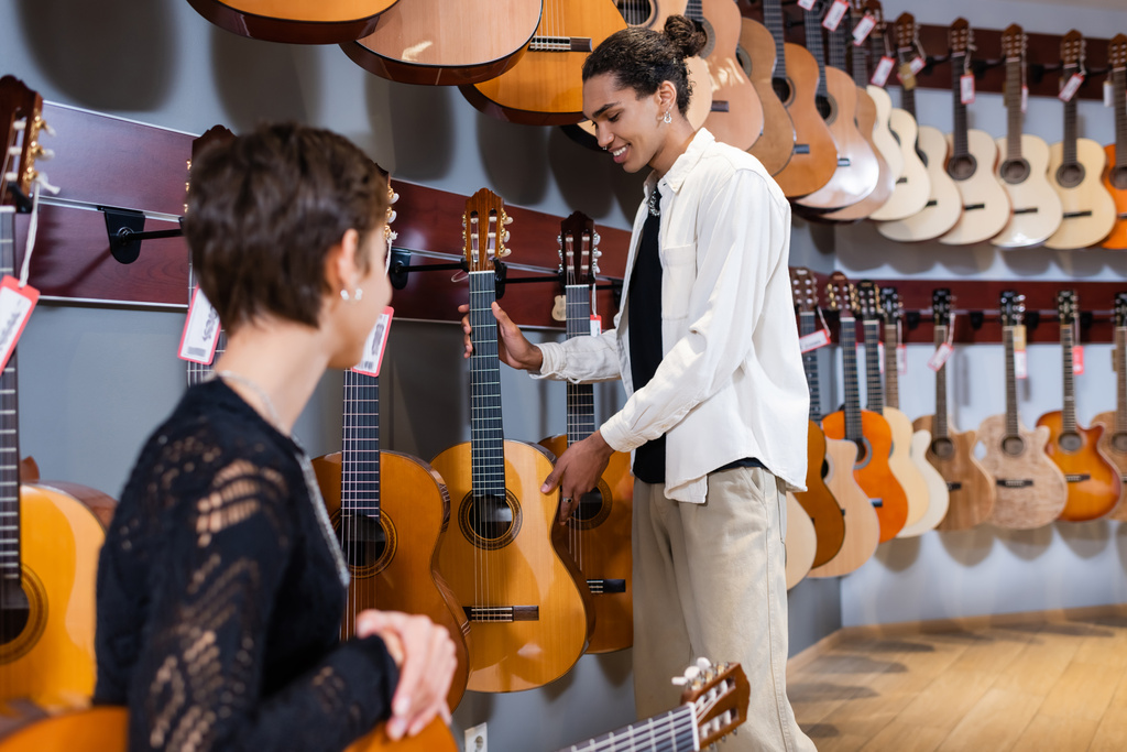 Positivo venditore afroamericano con chitarra acustica vicino al cliente offuscato nel negozio di musica strumentale  - Foto, immagini